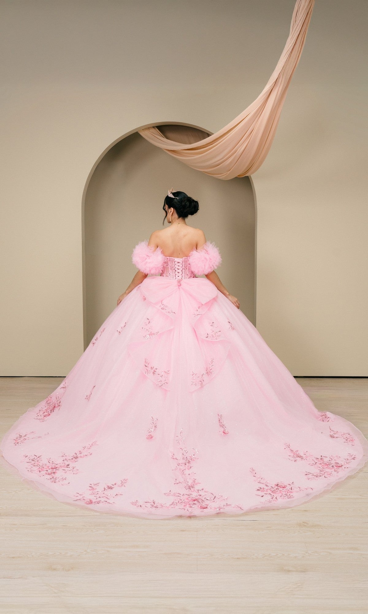 A woman wearing the Quinceanera Dress 8380J by Cinderella Couture, a pink ball gown with floral embroidery and a corset bodice, stands facing away from the camera before an arched backdrop with draped fabric.